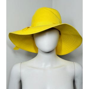May include: A bright yellow sun hat with a wide, flexible brim and a matching yellow ribbon band tied in a small bow on the left side. The hat features a rounded crown and a soft, unstructured fabric construction that drapes slightly over the brim.
