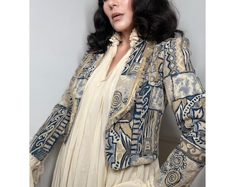 May include: A cropped jacket featuring a multi-colored tapestry pattern in shades of blue, beige, and cream. The jacket has long sleeves, a structured fit, and a frayed edge trim along the front opening. It is worn over a cream-colored dress.