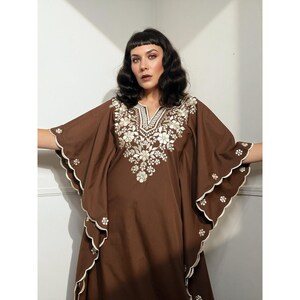 May include: A brown kaftan dress featuring cream-colored floral embroidery around the V-neckline and along the wide, scalloped sleeves. The edges of the sleeves are finished with cream piping and small embroidered flower accents throughout.