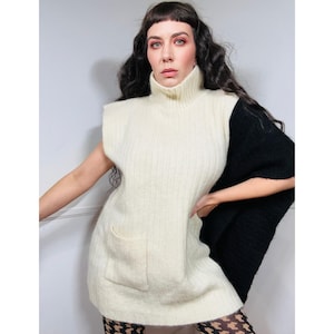 May include: A white and black knitted sweater vest with a high turtleneck and a patch pocket on the front. The sweater has a long, flowing sleeve on one side and a sleeveless design on the other.