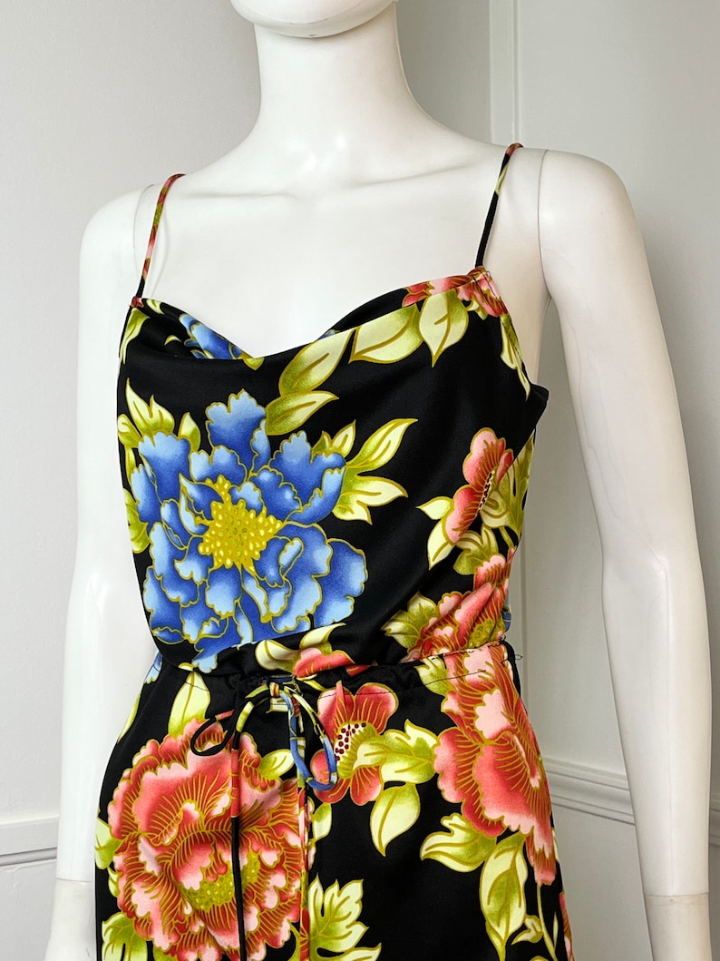 May include: A black sleeveless jumpsuit with a floral print featuring blue, pink, and yellow flowers. The jumpsuit has a cowl neck and a drawstring waist.