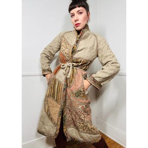 Puede incluir: Una larga gabardina beige de patchwork con cinturón de lazo. La gabardina está hecha de diferentes telas y patrones, incluyendo diseños florales, paisley y geométricos. La gabardina tiene un cuello alto y mangas largas.