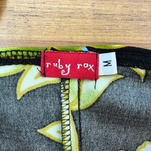 May include: A red tag with the text "ruby rox" in white lettering and a size "M" in white lettering on a black background. The tag is attached to a black fabric with a yellow and white floral pattern.