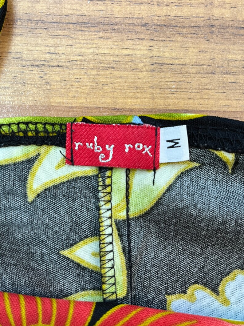 May include: A red tag with the text "ruby rox" in white lettering and a size "M" in white lettering on a black background. The tag is attached to a black fabric with a yellow and white floral pattern.