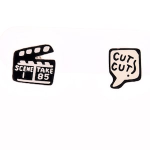 May include: A pair of film-themed earrings. One earring is a black and white movie clapperboard with the text "SCENE 1 TAKE 85." The other earring is a speech bubble with the words "CUT CUT!" in black.