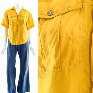May include: A short-sleeved, mustard yellow shirt with two chest pockets and button closures. The shirt is paired with blue denim, wide-leg pants. The outfit is displayed on a mannequin against a white background.
