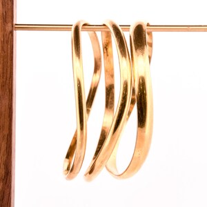 May include: Four gold-coloured metal bangles stacked on a wooden stand. The bangles are all different widths and have a slightly curved shape.