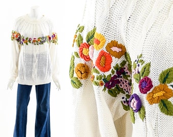 May include: Cream-colored sweater with colorful floral embroidery across the chest and sleeves. The sweater has a cable knit design on the front and is paired with blue denim jeans. The floral embroidery includes orange, yellow, purple, and green.