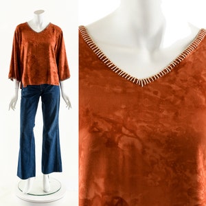 Puede incluir: Una blusa de manga larga de color óxido con cuello en V y mangas acampanadas. El escote y los bordes de las mangas están adornados con un diseño cosido en blanco y beige. La blusa se combina con unos vaqueros azules.