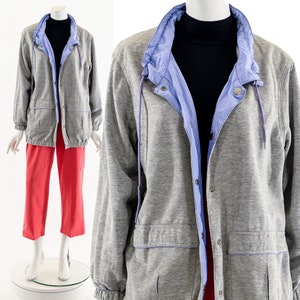 May include: A gray jacket with a light blue collar and lining, featuring a drawstring and snap closures. The jacket has two front pockets and is paired with red cropped pants. The outfit is displayed on a mannequin.