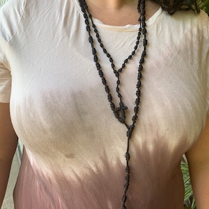 May include: A black knotted rosary necklace with a small cross at the end. The necklace is worn by a person with a light brown and white ombre t-shirt.