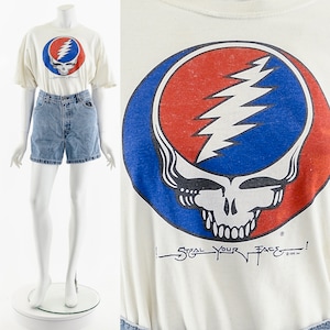 Puede incluir: Camiseta blanca con un logotipo de Grateful Dead Steal Your Face rojo, blanco y azul. El logotipo es una calavera con un rayo en la frente. El texto "Steal Your Face!" está escrito debajo del logotipo.