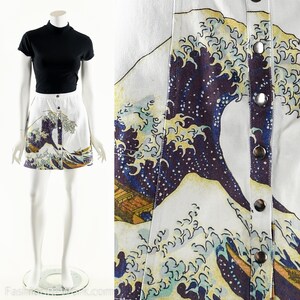 May include: A white denim mini skirt with a printed image of the Great Wave off Kanagawa by Katsushika Hokusai. The skirt has a button closure and a relaxed fit.