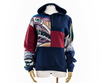 May include: A navy blue hoodie with a patchwork design featuring a green and gold football stadium scene, a red panel, and a blue and white pattern on the sleeves.