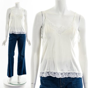Puede incluir: Un camisero blanco con delicados adornos de encaje en el escote y el dobladillo. La parte superior presenta finas correas de espagueti y un diseño de cuello en V. El camisero se combina con unos vaqueros acampanados azul oscuro, creando un look de inspiración vintage.