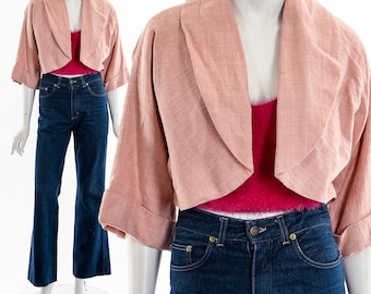 May include: A pale pink cropped jacket with a wide collar and rolled sleeves. The jacket is worn with a magenta top and dark blue jeans. The outfit is displayed on a mannequin.