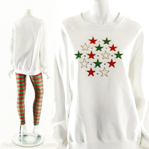 Puede incluir: Sudadera blanca con diseño de estrella. El diseño de estrella presenta estrellas rojas, verdes y doradas con detalles de pedrería. El conjunto incluye leggings a rayas rojas y verdes.