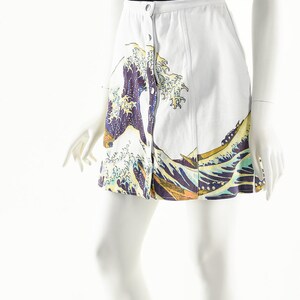 May include: A white skirt with a blue and yellow wave print and a button closure. The skirt is being modeled on a white mannequin.