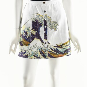 May include: A white skirt with a button-up front and a print of a large wave in blue, yellow, and purple. The skirt is paired with a black mock neck shirt.