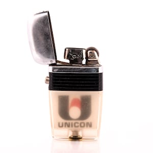 May include: A vintage UNICON lighter with a clear, rectangular body and a silver flip-top lid. The lighter features a black band and the UNICON logo in black with a red flame design. The text "VULCAN by Spain" is visible on the top.