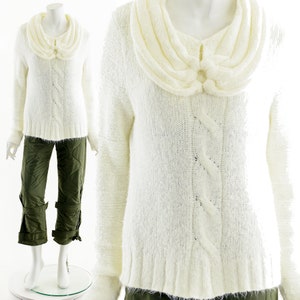 Può includere: Maglione color crema con motivo a trecce e ampio colletto ripiegato. Il maglione presenta un bottone decorativo sul colletto ed è abbinato a pantaloni verde oliva. L'outfit è esposto su un manichino.