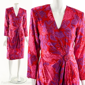 May include: A long-sleeved wrap dress with a V-neckline. The dress is made of a red and purple floral print fabric. The dress has a tie at the waist and a button detail.