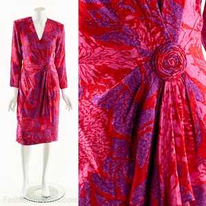 May include: A red and purple floral print dress with a long sleeve and a gathered waist. The dress has a large, fabric rose embellishment on the front.