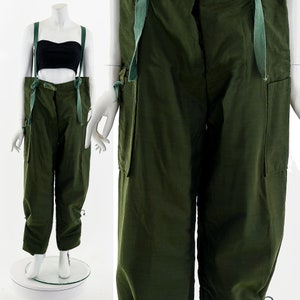 Könnte beinhalten: Grüne Cargohose mit Hosenträgern und großen Taschen. Die Hose hat einen Kordelzug am unteren Ende der Beine.