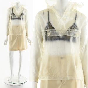 May include: A clear plastic raincoat with a hood and a button closure. The raincoat has a patch pocket on the front with the text "The  Knitter" printed on it.
