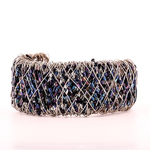 Op de afbeelding: Een zilverkleurige metalen manchetarmband met een geweven ontwerp. De armband is gevuld met kleine, rechthoekige, iriserende kralen in tinten blauw, paars en zwart. De manchet wordt op een witte achtergrond weergegeven.