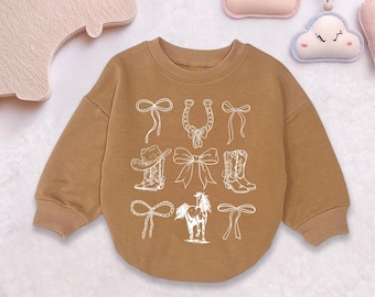 May include: A brown sweatshirt with a crew neck and long sleeves. The front of the sweatshirt features a white graphic design of cowboy-themed elements, including a cowboy hat, boots, bows, a horseshoe, and a horse.