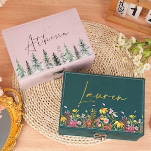 May include: Two personalized jewelry boxes. One is light pink with the name "Athena" and a forest design. The other is dark green with the name "Lauren" and a floral design. Both have metal clasps.