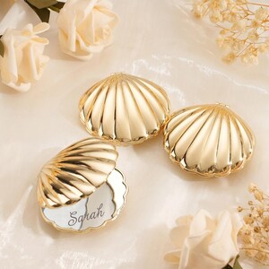 Pode incluir: Três espelhos de bolso em metal dourado em forma de concha. Dois espelhos estão fechados, enquanto um está aberto, revelando uma superfície refletora com o nome Sarah gravado em letra cursiva. As conchas apresentam um design canelado.