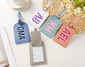 May include: A collection of personalized luggage tags in various colors, including blue, white, teal, and peach. Each tag features a unique three-letter initial combination in a contrasting color. The tags have a leather-like appearance and include a strap and buckle.