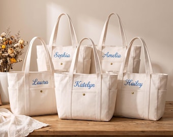 May include: Five off-white canvas tote bags with two top handles and a front pocket secured by a brass-colored snap button. Each bag features a unique name embroidered in blue cursive script: Sophia, Amelia, Laura, Katelyn, and Hailey.