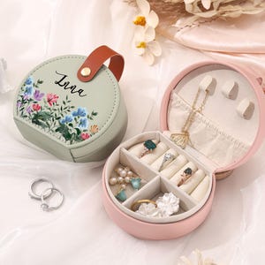 Puede incluir: Dos joyeros redondos, uno verde salvia y otro rosa, están abiertos sobre una superficie blanca. La caja verde tiene un diseño floral y el nombre "Irna" en escritura cursiva. La caja rosa está abierta, revelando compartimentos para anillos y collares. También se ve un par de anillos de plata.