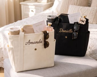 May include: Two canvas tote bags, one cream and one black, each featuring a front pocket with a snap button and a name embroidered in cursive script. The cream bag reads Amanda, and the black bag reads Danielle. Both contain tumblers and cards.