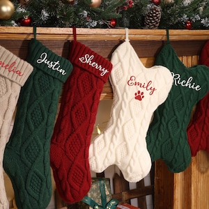 May include: Six cable-knit Christmas stockings in beige, green, and red hang from a wooden mantel. Each stocking is personalized with a name embroidered in a complementary color. One stocking has a paw print.