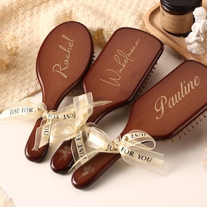 May include: Three personalized wooden hairbrushes with names engraved on them: "Rachel", "Winkelmann", and "Pauline". Each brush is tied with a cream ribbon that says "JUST FOR YOU". The brushes are brown with a dark handle.