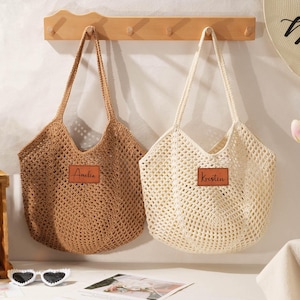 Puede incluir: Dos bolsos tote de tejido abierto, uno marrón y otro crema, cuelgan de un perchero de madera. Cada bolso tiene un parche rectangular de cuero con un nombre. Los bolsos tienen asas largas y están diseñados para llevar objetos.