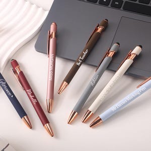Puede incluir: Una colección de bolígrafos personalizados en varios colores, incluyendo azul marino, burdeos, rosa, marrón, gris, blanco y azul claro. Cada bolígrafo presenta una punta y un clip dorados, con inscripciones de texto personalizadas. Los bolígrafos están colocados sobre una superficie blanca cerca de un portátil.