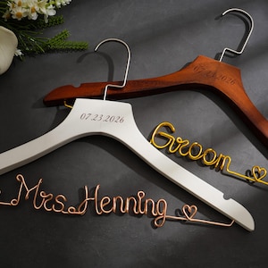 May include: Wedding hangers with custom details. One white hanger with "07.23.2026" and a copper wire "Mrs. Henning" sign. A brown hanger with "07.23.2026" and a gold wire "Groom" sign. Wedding decor.