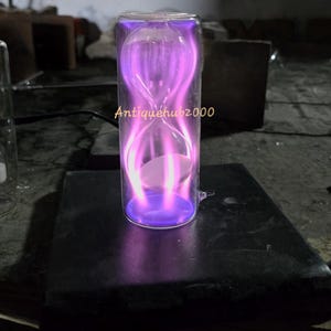 May include: A clear glass hourglass with a vibrant purple plasma effect. The hourglass shape is filled with a glowing, swirling light, and the text "Antiquehub2000" is visible on the glass. The base is a dark, rectangular shape.