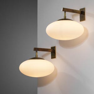 Puede incluir: Dos apliques de pared con una pantalla ovalada de vidrio esmerilado. Los apliques tienen un acabado de latón y un brazo de madera que se extiende desde la pared. El diseño es minimalista y moderno, adecuado para la iluminación interior.
