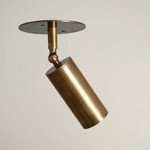 May include: A brass-colored, cylindrical spotlight fixture. The light is mounted on a round base and is angled downward. The fixture is designed for ceiling mounting and has a modern, minimalist aesthetic.