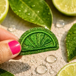 May include: A semi-circular embroidered patch shaped like a lime wedge, featuring vibrant green thread with detailed stitching to represent the fruit segments and rind, showcasing a textured surface and a bright, saturated green color palette.
