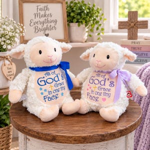 May include: Two plush white lamb toys with cream-colored faces and ears. The left lamb wears a blue ribbon and features blue embroidery reading All of God's Grace in one tiny Face. The right lamb wears a purple ribbon and matching purple text.