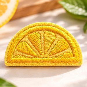 May include: A semi-circular embroidered patch featuring a bright yellow lemon slice design. The embroidery uses dense yellow thread to create the segments and rind of the citrus fruit, resulting in a textured, raised surface appearance.