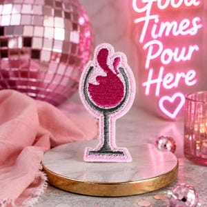 May include: An embroidered patch shaped like a wine glass filled with dark red liquid. The patch features a light pink border, a black stem and base, and a dark red wine graphic. The design is centered and stands upright on a circular display base.