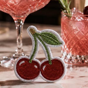 May include: An embroidered patch featuring two round red cherries with white highlights, connected by green stems and a single green leaf with gold thread detailing at the top, all outlined with a white border against a blurred background.